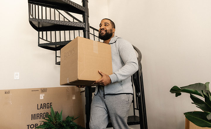 A smiling man by a staircase, walking and holding a cardboard moving box